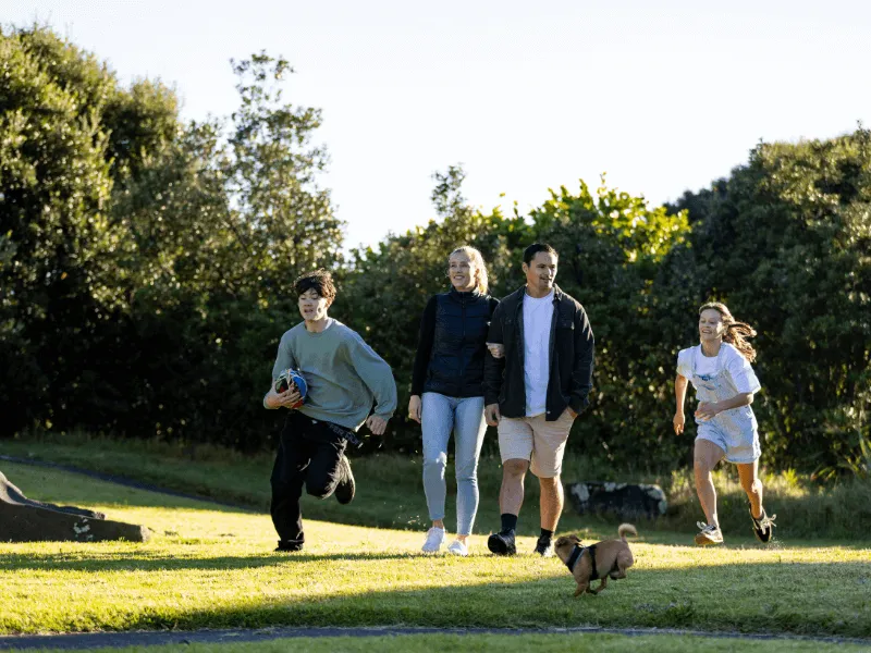 A family group, including two adults and three children, is enjoying a sunny day at the park. One child is running with a rugby ball, another is mid-run in casual clothes, and a small brown dog trots ahead on the grass. The adults walk together, smiling,