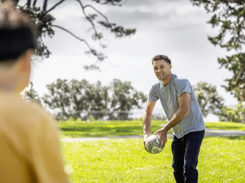 Two people playing rugby in a sunny park, one holding a rugby ball and smiling outdoors.