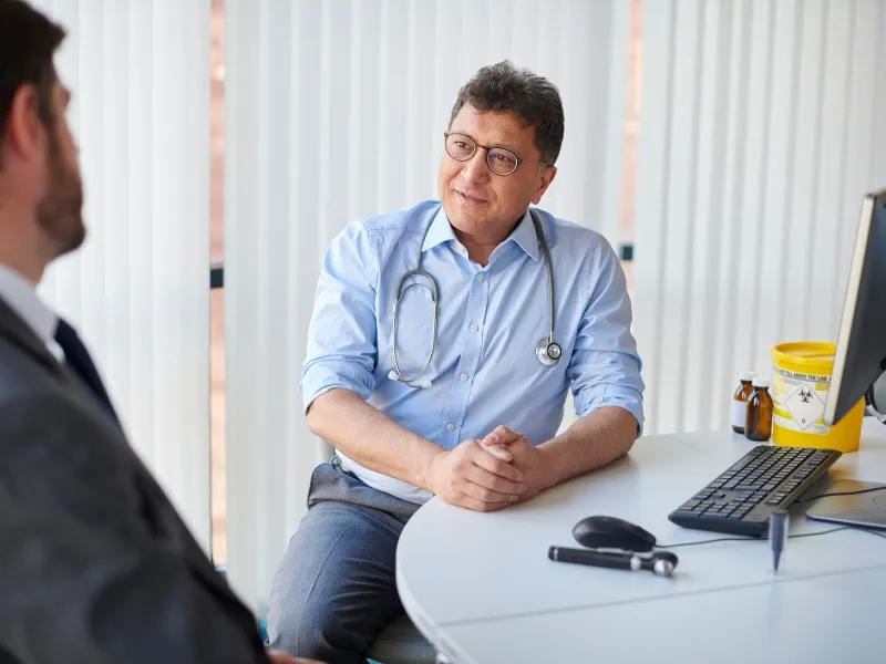 Middle‑aged man talking with doctor during heart health check, GP consultation in clinic office, discussing cardiovascular risk and heart disease prevention.