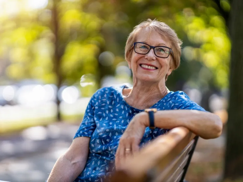 Smiling older woman sitting on park bench, enjoying healthy outdoor lifestyle and positive wellbeing after looking after her heart health.