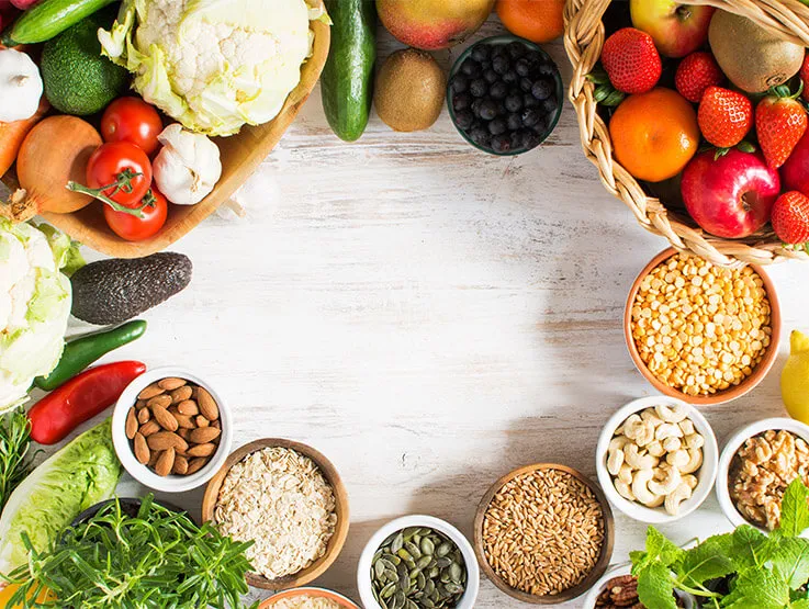 Variety of fruits and vegetables, cereals, and nuts on a white wooden table. Basket of strawberries, apples, oranges, kiwi, bowls of oats; broccoli