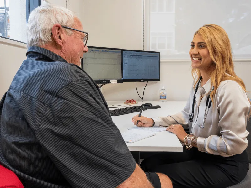 Doctor consulting with an older male patient in a modern medical office, discussing test results displayed on dual computer screens.