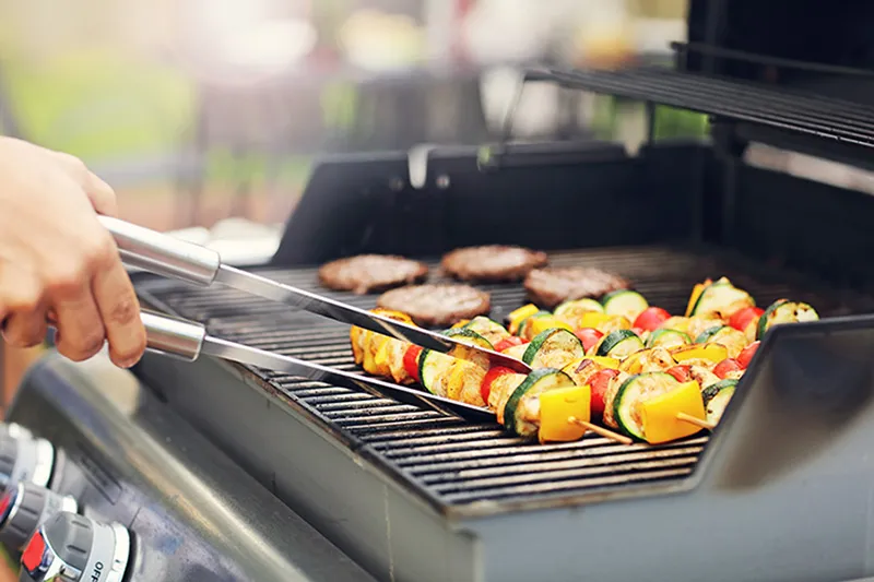 A hand holding a pair of tongs is turning vegetable kebabs on a BBQ grill. The kebabs comprise of colourful vegetables.