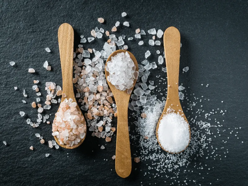 Three wooden spoons on a dark slate surface, each holding a different type of salt: pink Himalayan salt on the left, coarse sea salt in the center, and fine table salt on the right, with salt crystals scattered around each spoon.