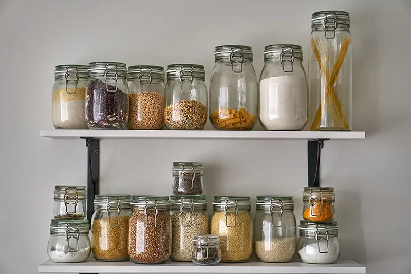Two shelves in a pantry hold glass jars containing bulk foods, such as oats, pasta, dried beans, rice.