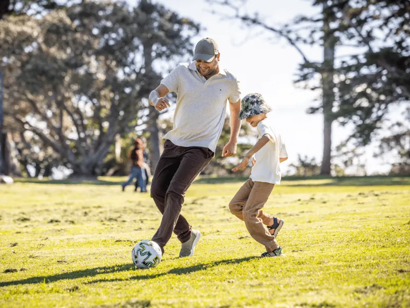 Adult and child playing football on grass in a sunny park, staying active together.