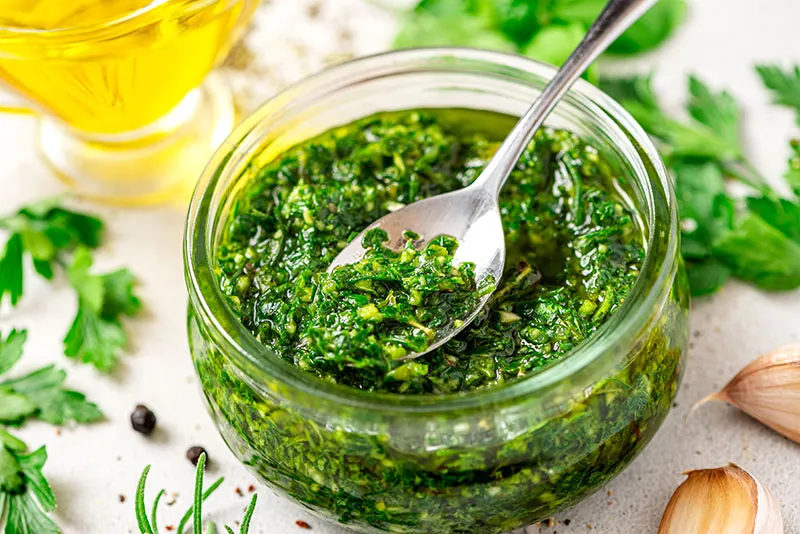 Chimichurri is a Latin American sauce made from various herbs, garlic and pepper. This image shows Chimichurri verde in a glass bowl on a white background. Sauce of parsley, oregano, rosemary, garlic, pepper, and oil.