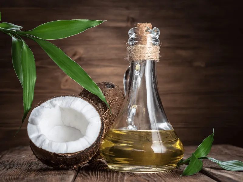 Glass bottle of coconut oil with fresh split coconut and green leaves on rustic wooden table.