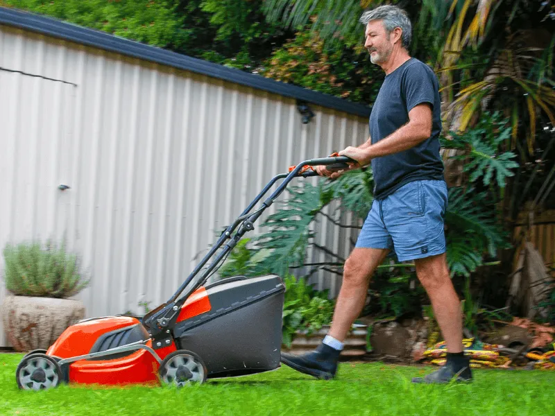 Middle-aged man mowing the lawn in a backyard with a bright orange electric lawnmower, wearing a black t-shirt and blue shorts, surrounded by lush greenery.