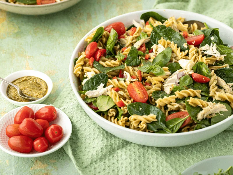 A large white bowl filled with pasta salad containing rotini pasta, shredded chicken, fresh spinach leaves, halved cherry tomatoes, and sliced cucumbers. Nearby, a small bowl of pesto dressing with a spoon and a plate of whole cherry tomatoes are set on a