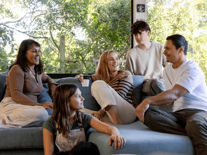 Six people, including adults and children, are sitting together on a light blue sectional couch in a living room with large windows and lush greenery outside. They are casually dressed, smiling, and engaged in conversation, creating a warm and relaxed fam