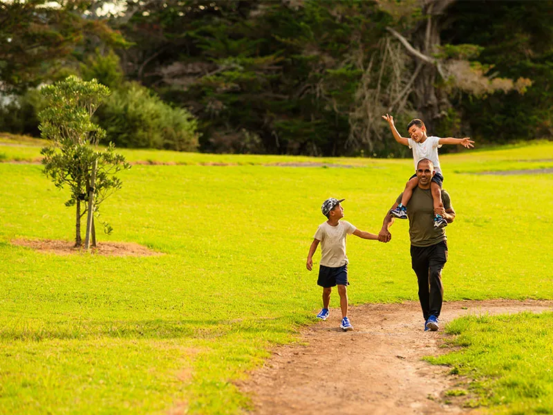 This image shows an adult male of Asian heritage, walking in a park with his two male children. He is holding the hand of a small boy, and has an even smaller boy riding on his shoulders. The boy on the shoulders has his arms raised above his head in joy.