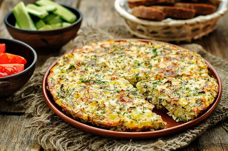 A frittata made with chicken, potatoes and vegetables depicted on a plate surrounded by tomatoes, cucumbers and sliced bread.