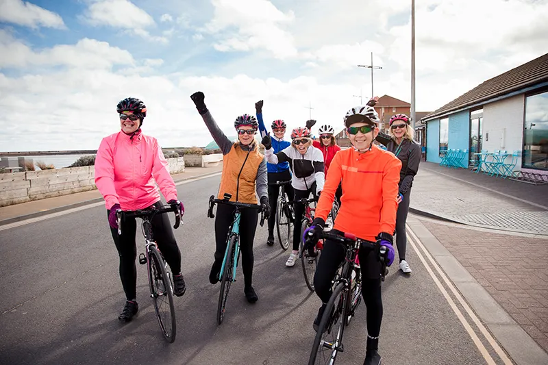 Group of older female cyclists enjoying a bike ride together in a small New Zealand town. They are grinning, and some are raising their hands in the air in celebration.