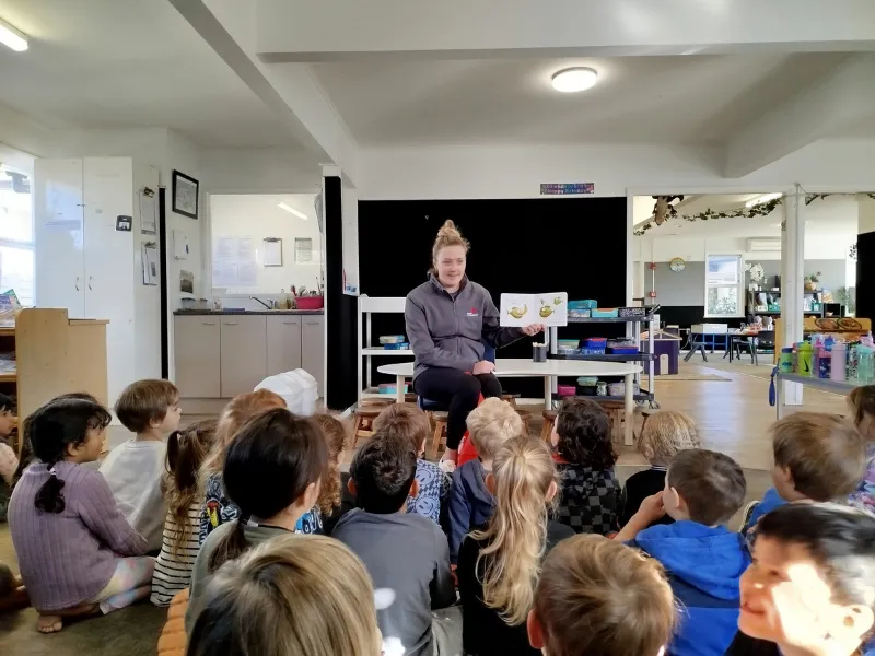 A group of young children sit on the floor of a classroom listening to an adult at the front who is holding up an illustrated book, leading a lesson about healthy food.