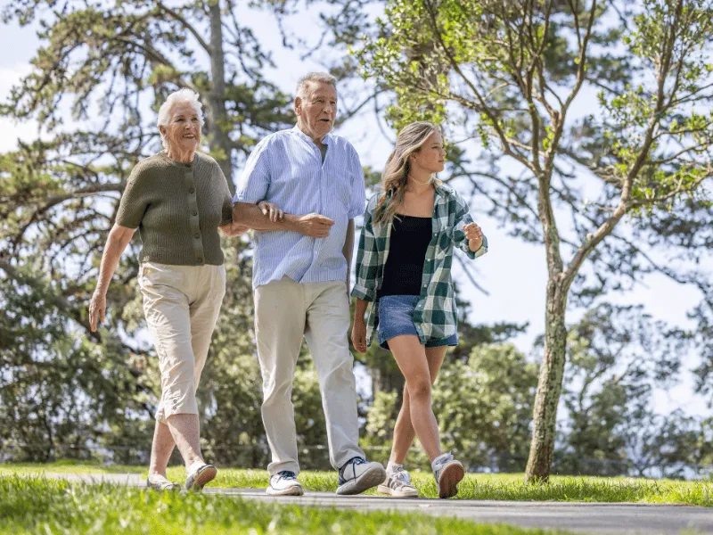 Two older adults and a younger woman walking together along a tree-lined park path.