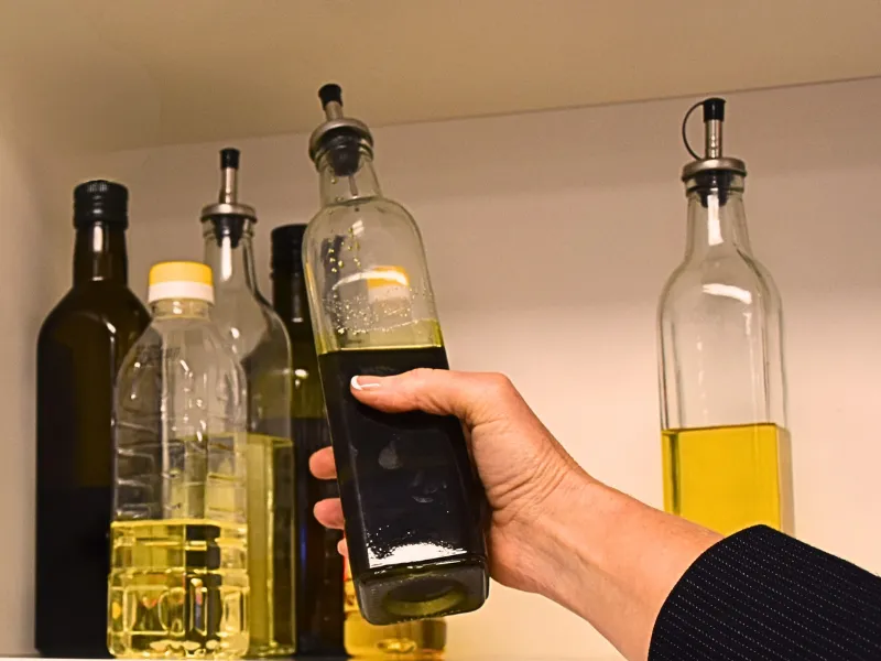 Hand reaching for a bottle of cooking oil from a pantry shelf filled with different vegetable and olive oil bottles.