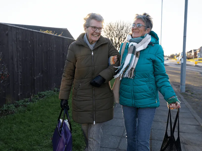 Two women wearing winter coats and scarves walk together on a sidewalk carrying shopping bags on a clear day.