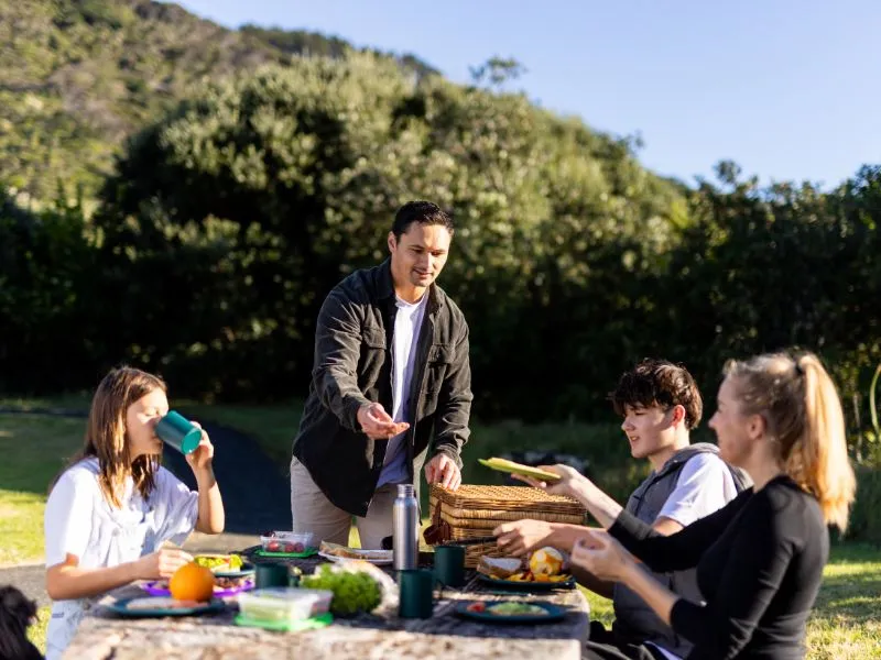 A family group of four is enjoying a picnic outdoors on a sunny day. They are gathered around a wooden table covered with colorful food and drinks. A man stands by a picnic basket, handing out items, while a woman and two teenagers sit, smiling and intera