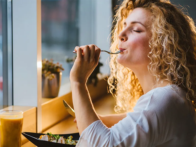 A young women sits at a table eating a healthy bowl of chicken salad. She has the fork in her mouth and her eyes are closed in an expression of enjoyment and satisfaction with her mouthful, as though she is savouring every flavour.