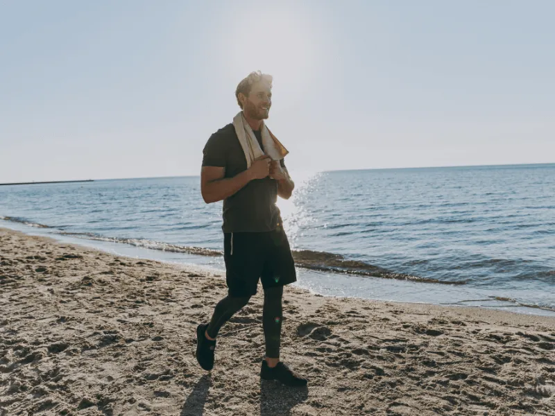 A person wearing athletic clothing walks along a sandy beach near the water’s edge, holding a towel around their neck, with the sun shining brightly overhead and the sea stretching into the distance.