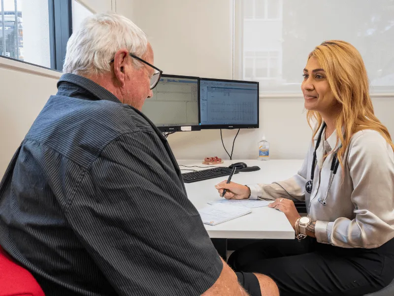 An older man sits across from a female doctor at a desk in a medical office. The doctor, wearing a stethoscope and business attire, writes notes on a paper while referencing information on two computer monitors. The office is bright with large windows and