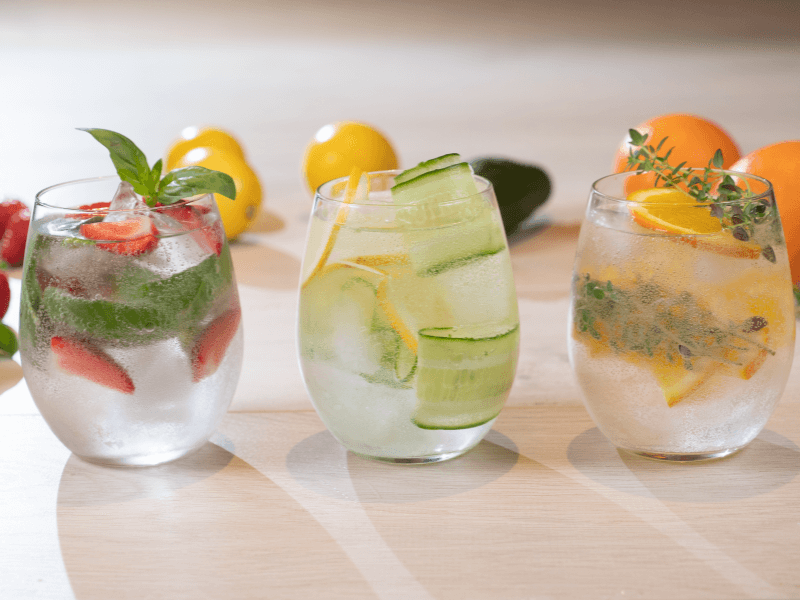 Three glasses of infused water on a light wooden surface: the left glass contains strawberries and basil, the middle glass has cucumber and lemon slices, and the right glass features orange slices and sprigs of fresh herbs, with whole lemons and oranges i