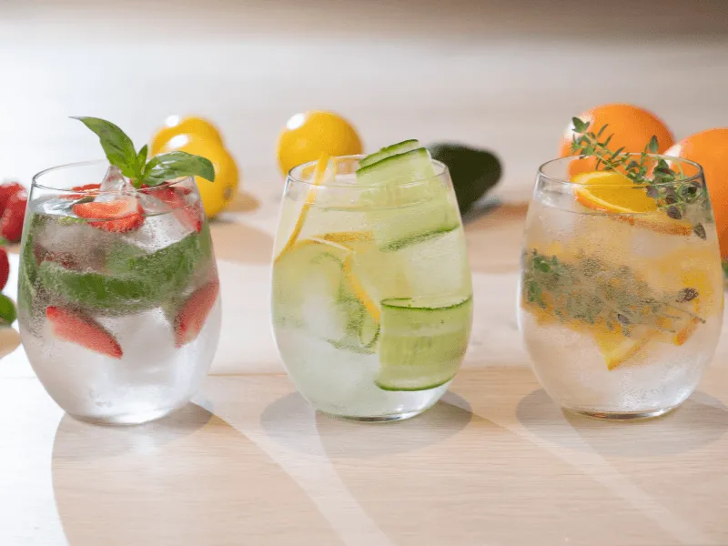 Three glasses of infused water on a light wooden surface: the left glass contains strawberries and basil, the middle glass has cucumber and lemon slices, and the right glass features orange slices and sprigs of fresh herbs, with whole lemons and oranges i