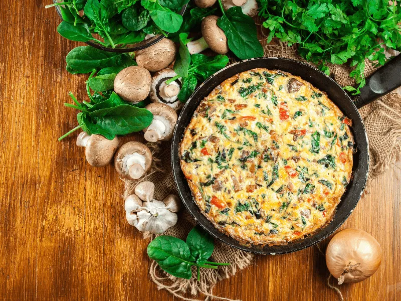 A freshly cooked vegetable frittata in a skillet surrounded by fresh ingredients including mushrooms, spinach, garlic, parsley, and an onion on a wooden table.