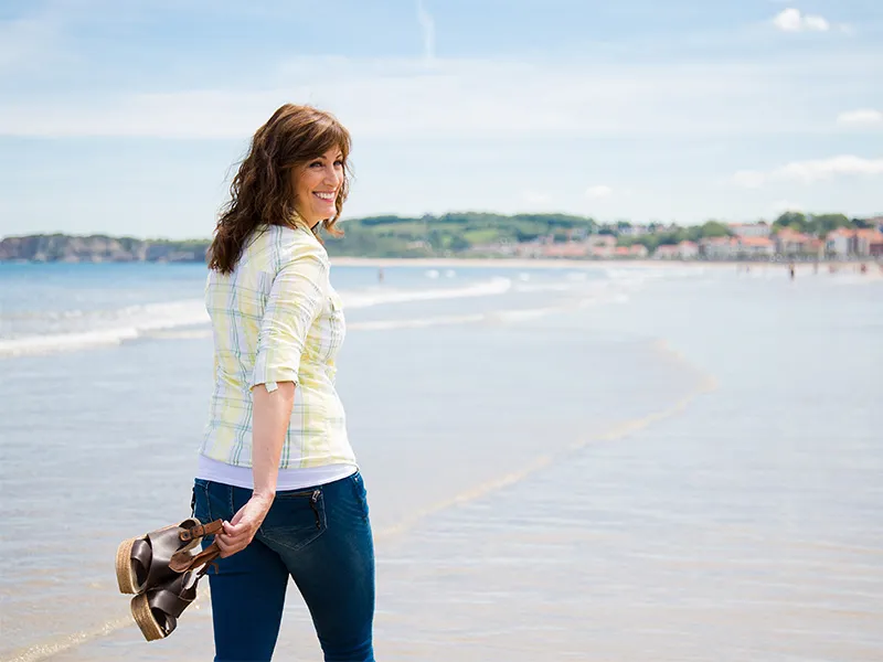 A middle aged women walking on a beach holding her sandals in her hand. She is looking over shoulder and smiling broadly into the camera.