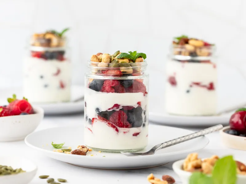 Glass jars filled with layers of Greek yoghurt, fresh mixed berries, nuts and seeds, healthy breakfast berry parfaits on white plates.