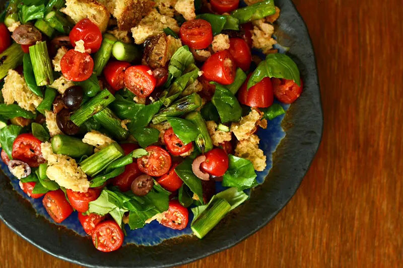 Large serving dish containing a panzella salad. Panzella means croutons. Other visible ingredients include asparagus, cherry tomatoes, spinach leaves.