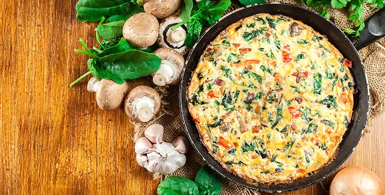 Frittata in pan on table surrounded by mushrooms and herbs