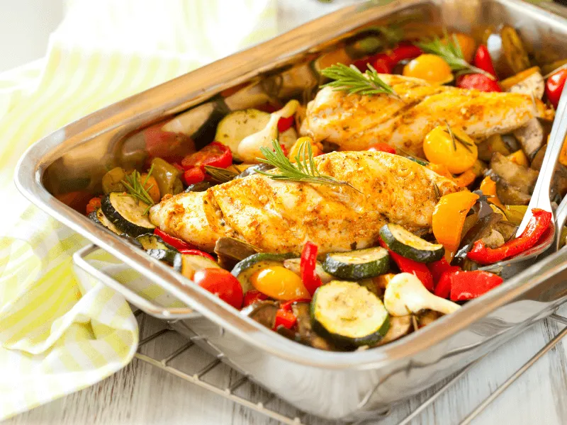 A stainless steel baking dish filled with roasted chicken breasts garnished with rosemary, surrounded by colorful roasted vegetables including zucchini, cherry tomatoes, red bell peppers, and onions. The dish is set on a cooling rack with a yellow and whi