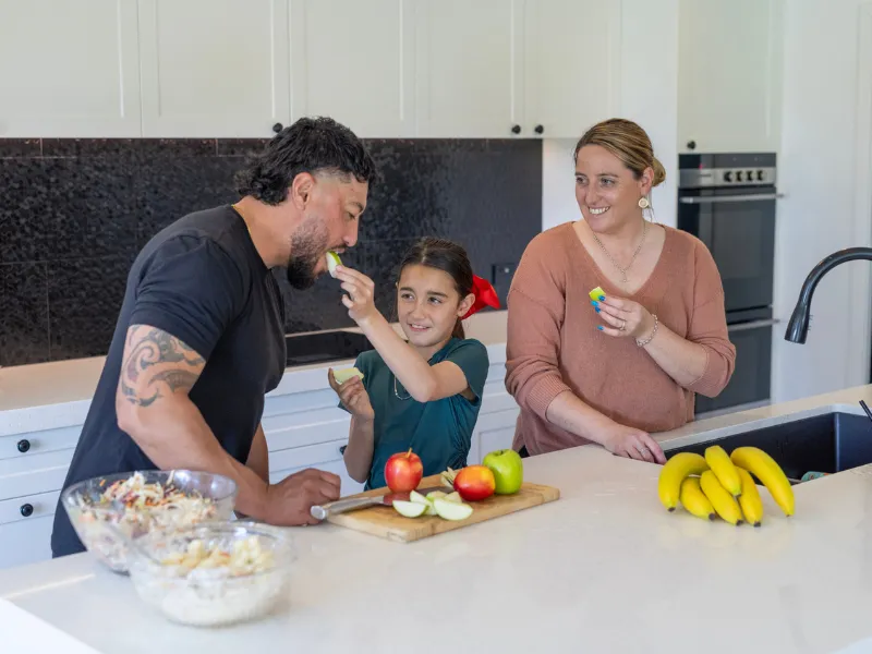 Smiling family in modern kitchen preparing a healthy snack, young girl feeding her dad apple slices while mum watches, fresh fruit on bench including apples and bananas.