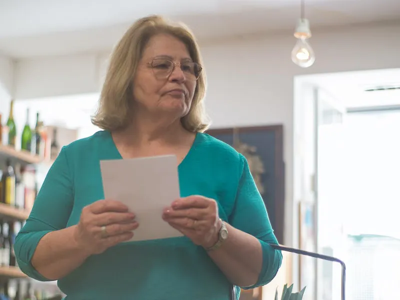 An older lady is shopping in a grocery store. She has a shopping basket over her arm and a shopping list in both hands. She wears a green t-shirt.