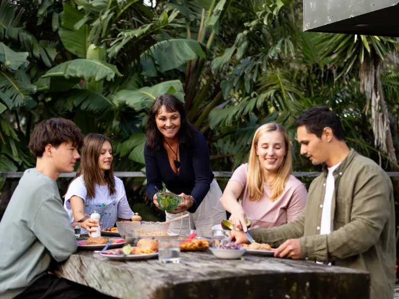 Group of friends enjoying a healthy summer BBQ with salads and grilled food around an outdoor wooden table surrounded by lush green native New Zealand bush.
