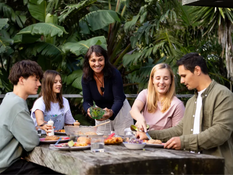 Group of people enjoying a healthy outdoor barbecue, sharing salads and colourful dishes together around a rustic wooden table in a lush garden setting.