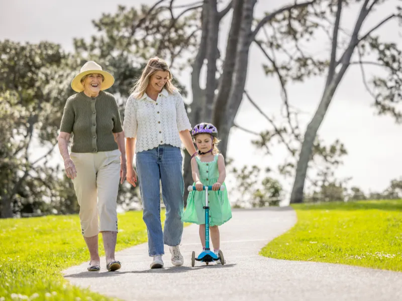 Three generations family walking outdoors on a park path, grandmother and mum beside young girl riding a scooter with helmet, active healthy lifestyle.