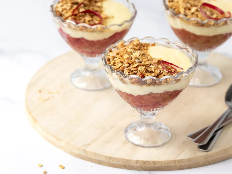 Individual dessert glasses filled with layered rhubarb, creamy custard and crunchy granola topping, healthy fruit crumble parfait on wooden board