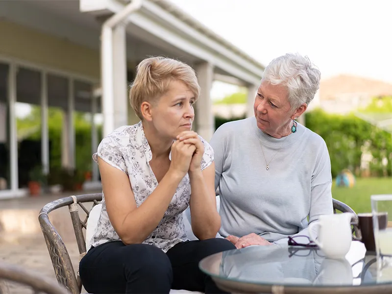 In this image we see two adult women - a mother and her daughter - sitting outside at a garden table. The daughter looks visibly stressed and upset and the mother is rubbing her back in comfort.