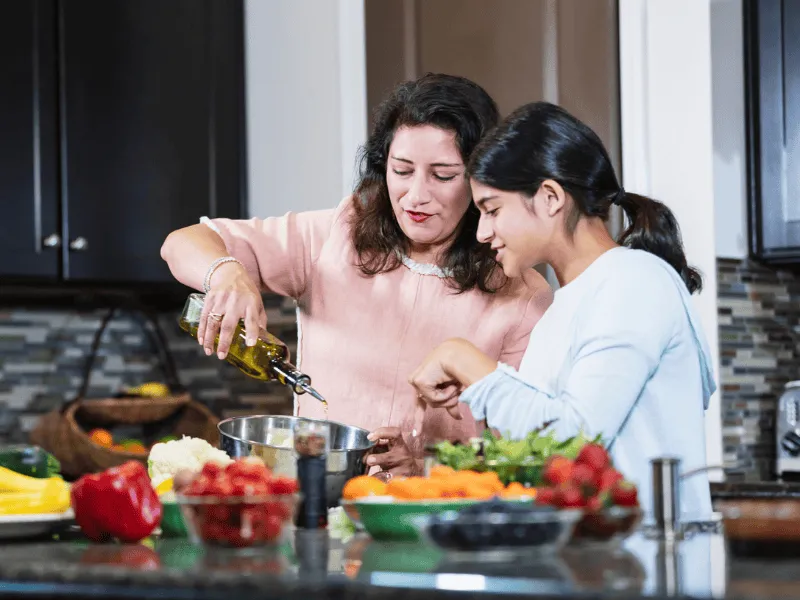 Two people cooking together in a kitchen, pouring olive oil over a bowl of fresh vegetables and salads.