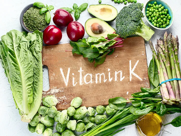 A selection of vitamin k-rich vegetables is displayed around a chopping board. The vegetables include romaine lettuce, basil, plums, avocado, broccoli, peas, asparagus, spinach, Brussel sprouts.