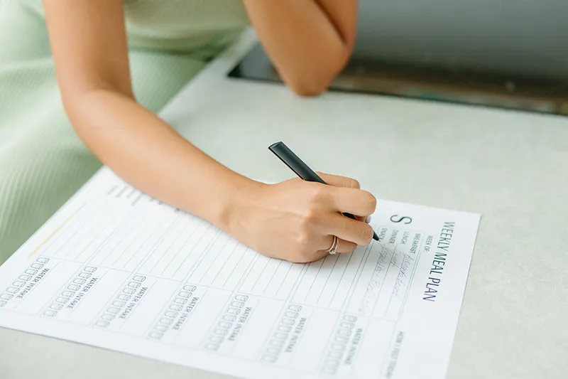 Image shows a female hand writing down some options in a weekly meal planner. The title Weekly meal planner is visible at the top of the page. Also visible are separate fields for breakfast, lunch and dinner. 