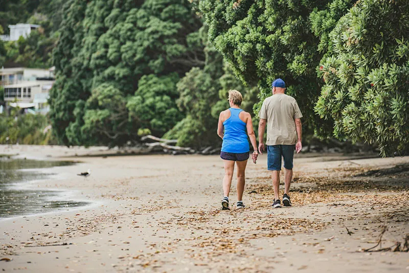 A senior man and women are walking along a New Zealand beach. They have their backs to the camera as they walk away from us. 