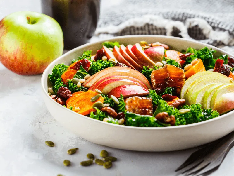 A fresh salad in a white bowl featuring sliced red and green apples, kale, roasted vegetables, nuts, seeds, and dried fruits. An apple and a drink are visible in the background, and a fork is placed nearby on the light-colored table.
