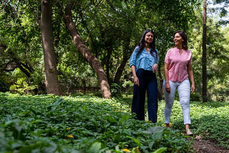 Two women walk through a park with dense foliage, while chatting with one another.