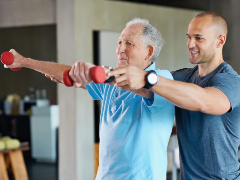 Physiotherapist supporting older man with arm exercises using light dumbbells during rehabilitation session in a gym or clinic setting.