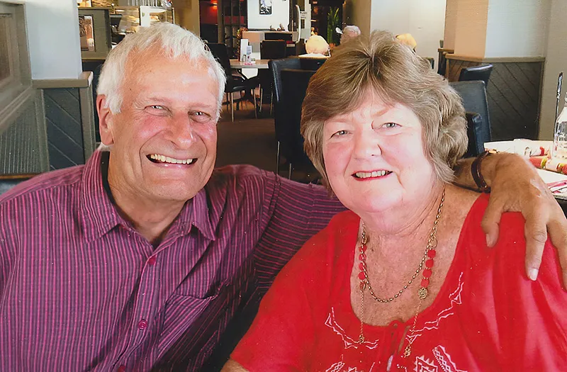 Barry Eastwick and his wife, are shown sitting at a dining table. There are Christmas decorations on the table. The couple are smiling into the camera and Barry has his arm around his wife's shoulders.  