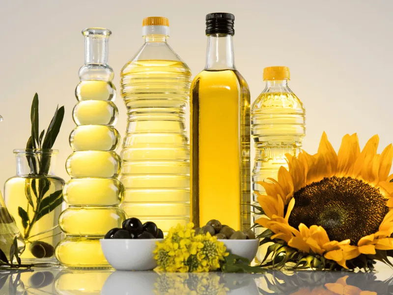 Assorted bottles of vegetable and olive oil with olives and sunflower displayed on a reflective kitchen surface.